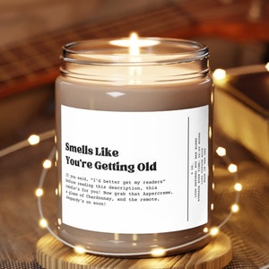 May include: A brown candle with a white label that reads "Smells Like You're Getting Old". The label has a humorous description about getting older. The candle is sitting on a wooden coaster and surrounded by white fairy lights.