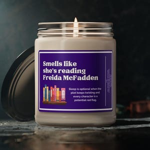 May include: A scented candle in a glass jar with a silver lid. The candle has a purple label with the text "Smells like she's reading Freida McFadden". The label also includes a graphic of books and a quote about sleep.