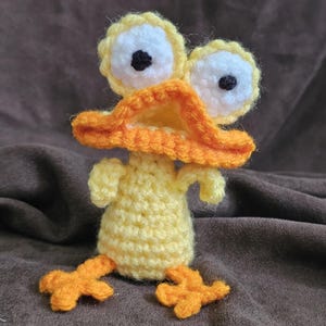 May include: A handmade crochet duck with large, white eyes with black pupils, a bright orange beak, and matching feet. The duck is primarily yellow, with orange accents. The craftwork is displayed against a brown background.
