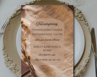 May include: A Friendsgiving invitation on a white plate, set on a decorative gold-colored charger. The invitation features text in a soft brown arch, including "Friendsgiving," "Thanksgiving Dinner," and the date and time. Dried flowers and a knife are also visible.