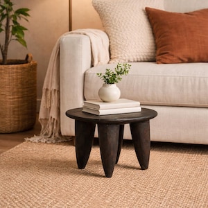 May include: A small, dark brown, three-legged wooden side table with a round top. The table holds two books and a small white vase with greenery. The table sits on a woven jute rug in front of a cream-colored sofa.