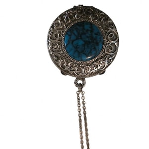May include: A round silver-toned pendant featuring an intricate, embossed scrollwork border surrounding a central, circular blue stone with dark veining. A delicate, thin silver-toned chain is attached to the bottom of the circular pendant.