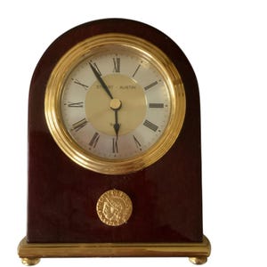 May include: A gold-toned clock with Roman numeral markings on a dark wood base. The clock face reads "Stuart Austin Quartz".