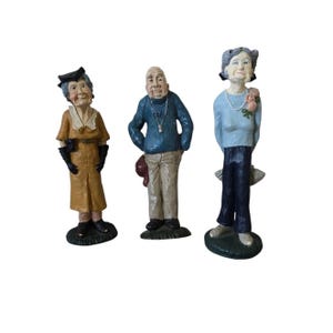 May include: Three ceramic figurines depicting elderly people. The figures are dressed in various outfits, including a mustard-colored coat, a blue sweater, and a light blue top. Each figure stands on a small, green base. The figurines have detailed facial features and are painted with realistic colors.