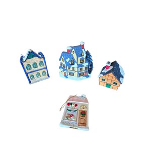 Könnte beinhalten: Vier Keramik-Weihnachtsdorfhäuser. Die Häuser haben schneebedeckte Dächer und sind in verschiedenen Farben bemalt, darunter Blau, Gelb und Braun. Ein Haus hat einen kleinen Weihnachtsbaum daneben. Die Häuser stehen auf weißem Hintergrund.