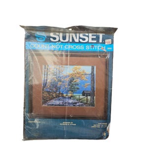 Op de afbeelding: Een Sunset Count-Not kruissteekpakket met een ingelijste landschapsscène van een landweg. Het kunstwerk toont een beek, bomen met gele en groene bladeren en een klein gebouw. De kit is verpakt in een doorzichtige plastic zak.