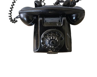 Pode incluir: Um telefone de disco preto com um cabo espiral. O telefone tem um receptor preto e um disco preto com números brancos.