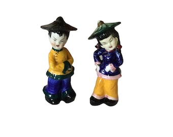 Könnte beinhalten: Zwei kleine Keramikfiguren von Menschen in traditioneller chinesischer Kleidung. Die Figuren sind in leuchtenden Farben bemalt, darunter Gelb, Blau, Grün und Pink. Die männliche Figur trägt ein gelbes und blaues Outfit mit einem schwarzen Hut, während die weibliche Figur ein blaues und gelbes Outfit mit einem grünen Hut trägt.