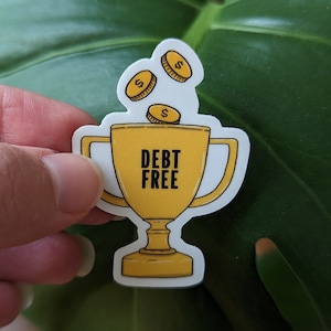 May include: A white sticker with a yellow trophy illustration. The trophy has the words "DEBT FREE" on it. Three gold coins with dollar signs are falling into the trophy.