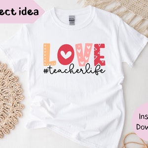 Pu&ograve; includere: Maglietta bianca con la parola "LOVE" in lettere rosse, rosa e arancioni con cuori e altri motivi. Il testo "#teacherlife" &egrave; sotto la parola "LOVE".