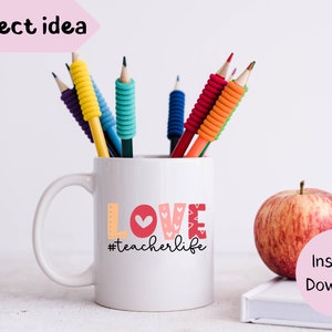 Pu&ograve; includere: Una tazza in ceramica bianca con una grafica "LOVE" rossa, rosa e bianca e il testo "#teacherlife" in nero. La tazza &egrave; appoggiata su una superficie bianca con una mela rossa e matite in una tazza.