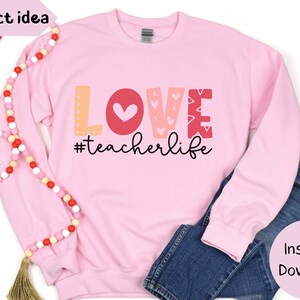 Pu&ograve; includere: Felpa rosa con la parola "LOVE" in lettere colorate e un design a forma di cuore. Il testo "#teacherlife" &egrave; sotto la parola "LOVE".