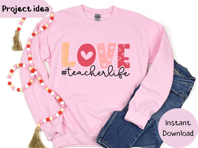 Pu&ograve; includere: Felpa rosa con la parola "LOVE" in lettere colorate e un design a forma di cuore. Il testo "#teacherlife" &egrave; sotto la parola "LOVE".