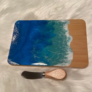 May include: A rectangular wooden serving board with a blue, green, and white epoxy resin ocean wave design. The board comes with a small wooden handled butter knife.