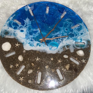May include: A round wall clock featuring a resin design of a beach scene with brown sand, white sea foam, and blue ocean water. The clock face includes embedded seashells, starfish, and twelve rectangular cutouts for hour markers with brown hands.
