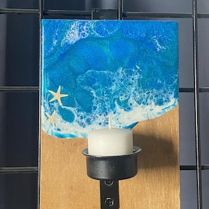 May include: A decorative wall-mounted candle holder. The top section features a blue and white resin design resembling ocean waves, with three starfish accents. A black metal candle holder is attached to a wooden base, holding a white candle.