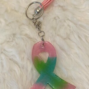 Pu&ograve; includere: Un portachiavi a forma di nastro in resina rosa e verde con glitter e un fiocco rosa.