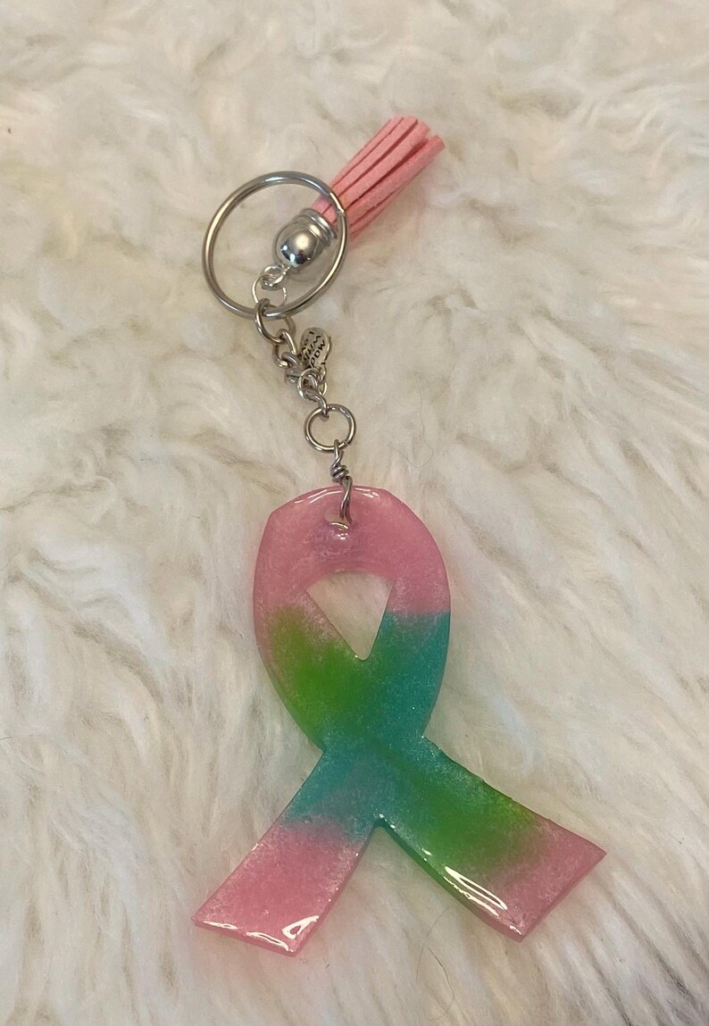 Pu&ograve; includere: Un portachiavi a forma di nastro in resina rosa e verde con glitter e un fiocco rosa.