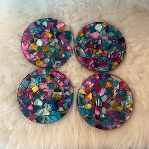Könnte beinhalten: Vier runde Untersetzer mit einem Mosaikdesign. Die Untersetzer haben eine klare Harzbasis, die mit farbenfrohen, irisierenden Muschelteilen in Blau-, Rosa-, Lila- und Gelbtönen gefüllt ist. Sie sind auf einer weißen, flauschigen Oberfläche angeordnet.