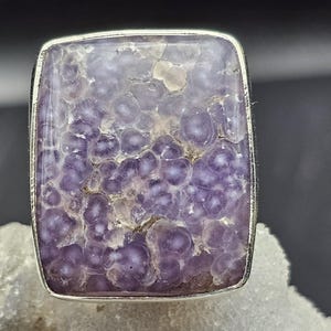 Op de afbeelding: Een vierkante ring met een zilveren zetting en een grote, getextureerde paarse steen. De steen heeft een bubbelpatroon in tinten lavendel en wit. De ring staat tegen een donkere achtergrond.