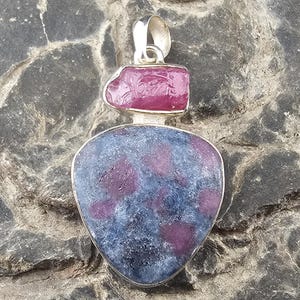 May include: A silver pendant with a large, shield-shaped blue and pink gemstone, and a smaller, rectangular pink stone above. The pendant has a silver loop for a chain. The stones have a natural, textured appearance.