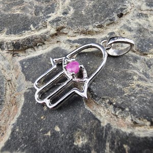 Puede incluir: Colgante de mano de Hamsa plateado con una gema rosa brillante en el centro. El colgante tiene un acabado pulido y un lazo para una cadena. El fondo es una piedra gris texturizada.