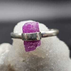 May include: A silver ring featuring a vibrant, raw, fuchsia-colored gemstone. The gemstone is rectangular and held in place by a silver band. The ring is displayed on a white, textured surface, with a blurred background.