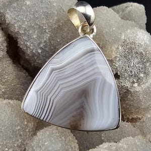 May include: A triangular pendant with a silver bail. The pendant features a polished, banded agate stone in shades of grey and white. The stone is set in a silver bezel, creating a striking contrast. The pendant is likely a piece of jewellery.