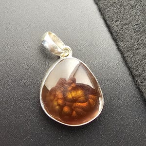 May include: A teardrop-shaped fire agate pendant set in a polished silver bezel. The gemstone features a translucent brown body with a vibrant, iridescent orange and gold botryoidal inclusion visible within the center of the stone.