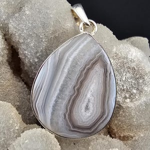 May include: A silver pendant with a teardrop-shaped agate stone. The stone displays bands of grey, white, and brown. The pendant has a silver bail. The background is a textured, light-coloured surface, possibly quartz.