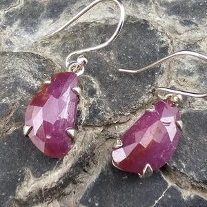 May include: Silver drop earrings with faceted, teardrop-shaped, deep red gemstones. The gemstones are secured by small silver prongs, and the earrings have a simple hook design.