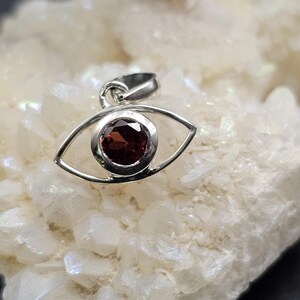 Może przedstawiać: Srebrny wisiorek w kształcie oka z czerwonym kamieniem szlachetnym w środku. Wisiorek leży na białym tle.