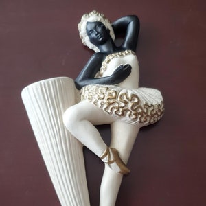 Peut inclure: Vase mural décoratif en forme de ballerine. La figurine a la peau noire, les cheveux blancs et porte un tutu blanc avec des détails dorés. Le vase blanc présente un design cannelé vertical.
