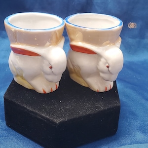 May include: Two white ceramic egg cups shaped like rabbits with red and blue accents. The cups are sitting on a black hexagonal surface.