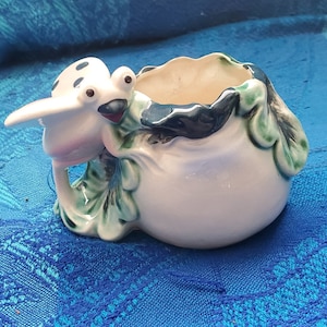May include: A white ceramic planter shaped like a ladybug with green and blue leaves. The planter has a small opening at the top for planting.