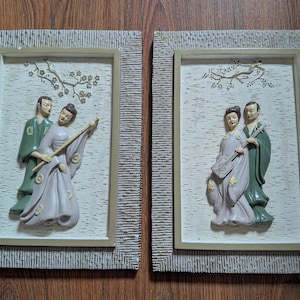 May include: Two framed wall art pieces featuring Asian-inspired relief sculptures. Each frame has a textured, light-colored border. The sculptures depict figures in traditional attire, one with a long instrument and the other with a stringed instrument.