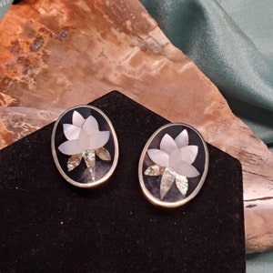 May include: A pair of oval-shaped earrings with a black background and a white flower design made of mother of pearl. The earrings have a silver-toned metal frame.