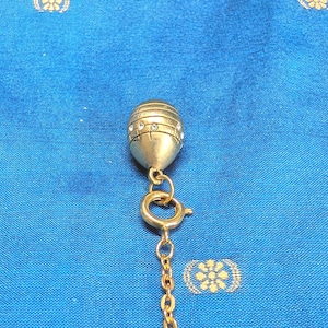 May include: A gold pendant necklace featuring a teardrop-shaped charm. The charm is decorated with small, clear crystals and horizontal lines. The necklace has a fine chain and a clasp. The background is a blue fabric with a floral pattern.