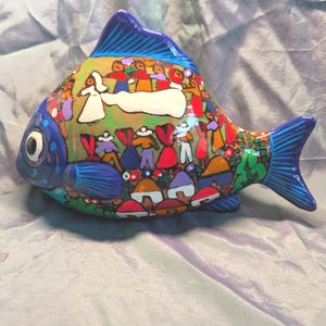 May include: A ceramic fish ornament, brightly painted with a folk art design. The fish has a blue head and tail, and a colourful body depicting a village scene with figures in various outfits. The artwork includes houses, trees, and a wedding scene.