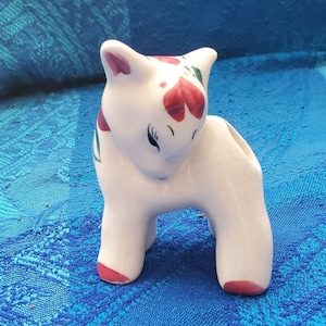 Pode incluir: Uma figura de cerâmica branca de um animal estilizado, possivelmente um gato ou um veado, com detalhes florais vermelhos e verdes. A figura tem um corpo curvo, com detalhes vermelhos nas orelhas e nos pés. Os olhos estão fechados, sobre um fundo azul.