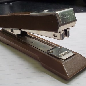 May include: A brown metal stapler with a silver top. The stapler has the text "APSCO 2004" on the top.