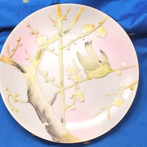May include: A decorative plate with a pink and white background featuring a gold-painted tree with a bird perched on a branch. The tree has delicate flowers and leaves.
