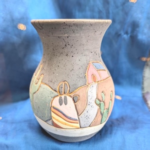 May include: A speckled grey ceramic vase featuring a hand-painted desert landscape scene. The design includes a pink house, a winding path, and green cacti against a tan and grey background. The vase has a flared rim and a white band at the base.