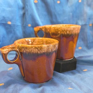 Può includere: Due tazze in ceramica con una smaltatura marrone lucida e un caratteristico motivo a goccia color crema attorno al bordo superiore. Ogni tazza presenta un manico robusto con apertura circolare, una finitura liscia e riflettente e forma conica.