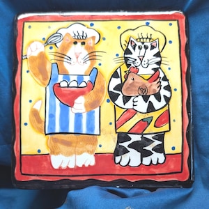 May include: Square ceramic plate with a design of two cartoon cats in chef outfits. One cat holds a whisk and eggs, the other a chicken. The plate has a yellow background with blue dots, a red border, and a black edge.