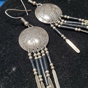 Könnte beinhalten: Silberne Ohrringe mit einem runden Anhänger mit einem indianischen Design. Der Anhänger ist an einer Kette aus silbernen und schwarzen Perlen befestigt.