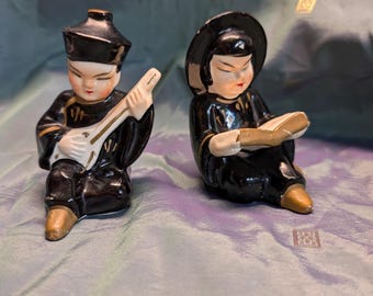 Könnte beinhalten: Zwei Keramikfiguren, die asiatische Figuren darstellen. Eine Figur spielt ein Saiteninstrument, die andere liest ein Buch. Beide tragen schwarze Roben mit goldenen Akzenten und bemalten Gesichtern. Die Figuren haben goldfarbene Füße.