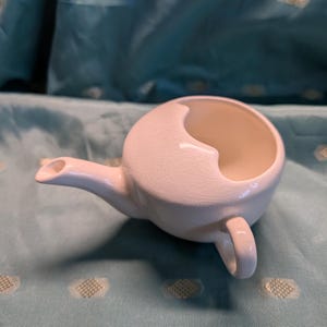 May include: A white ceramic teapot with a unique crescent-shaped opening and a small handle. The teapot has a spout and a crackled glaze finish, suggesting a vintage or handcrafted aesthetic. The design is simple yet distinctive.