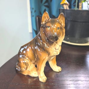 Könnte beinhalten: Eine Keramikfigur eines braunen und schwarzen Deutschen Schäferhundes, der sitzt und den Kopf leicht zur Seite geneigt hat. Der Hund hat eine glänzende Oberfläche und realistische Details.