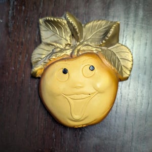 Puede incluir: Una manzana de cerámica amarilla con una cara sonriente y una lengua que sobresale. La manzana tiene un diseño de hojas doradas en la parte superior.